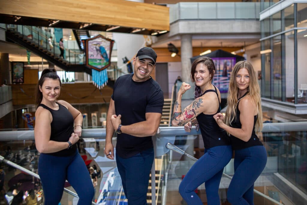Four personal trainers posing confidently in athletic wear, showcasing strength and fitness, with a modern training facility in the background, emphasizing RxFit's focus on personalized at-home training for seniors.