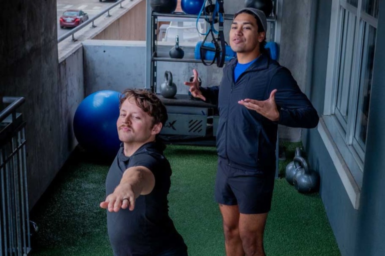 Two men demonstrating fitness poses on a balcony gym, showcasing wellness culture in Austin, with exercise equipment in the background.