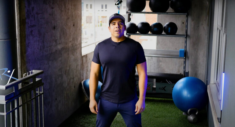 Fitness expert in casual workout attire standing in a home gym setting with exercise equipment, promoting at-home functional fitness routines for busy professionals.