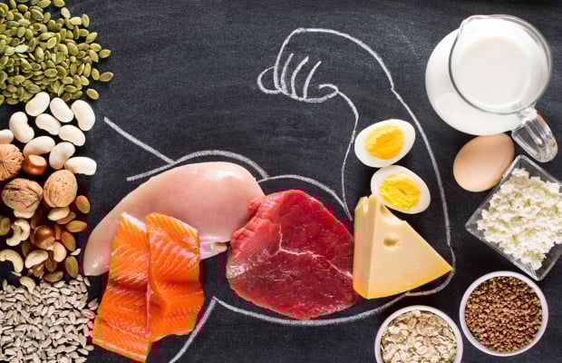 A variety of high-protein foods including salmon, chicken, beef, cheese, eggs, milk, seeds, and legumes arranged in front of a chalk drawing of a flexing arm.