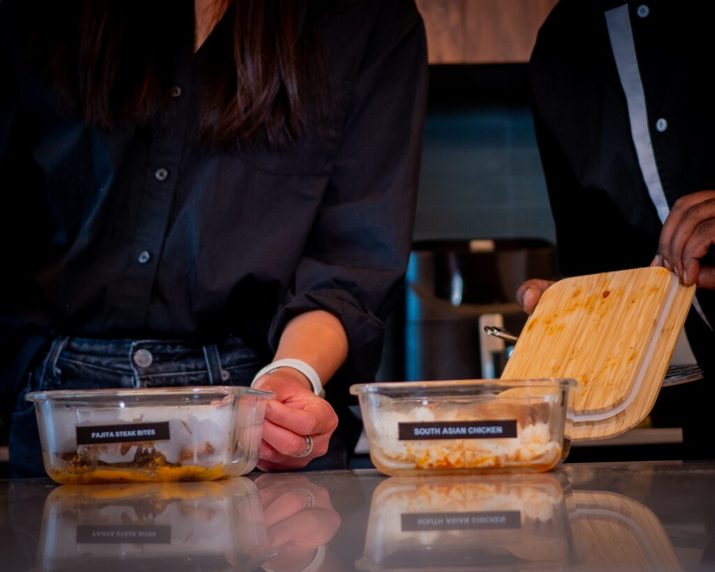 Person holding container labeled "Fajita Steak Bites" and another person preparing "South Asian Chicken" meal prep dishes, showcasing balanced meal options from RxFit for nutritional health.