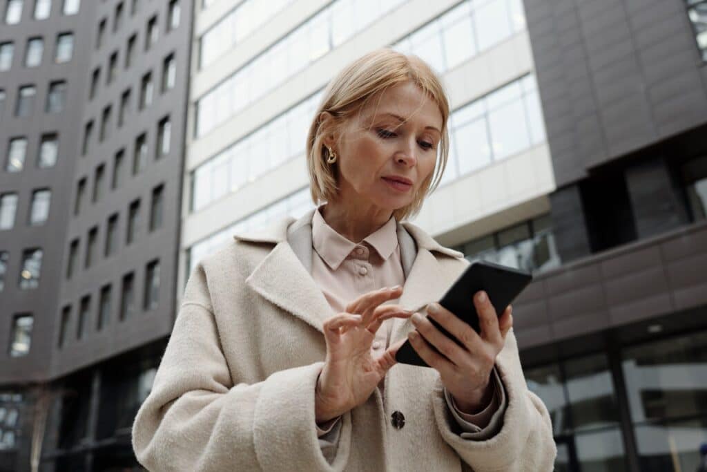 Woman in a beige coat using smartphone outdoors, surrounded by modern buildings, reflecting a focus on communication and personal engagement for injury recovery services.
