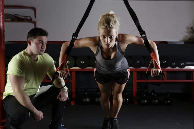 Woman performing TRX suspension training exercise with male trainer observing, gym setting with weights in background, emphasizing fitness and muscle growth.