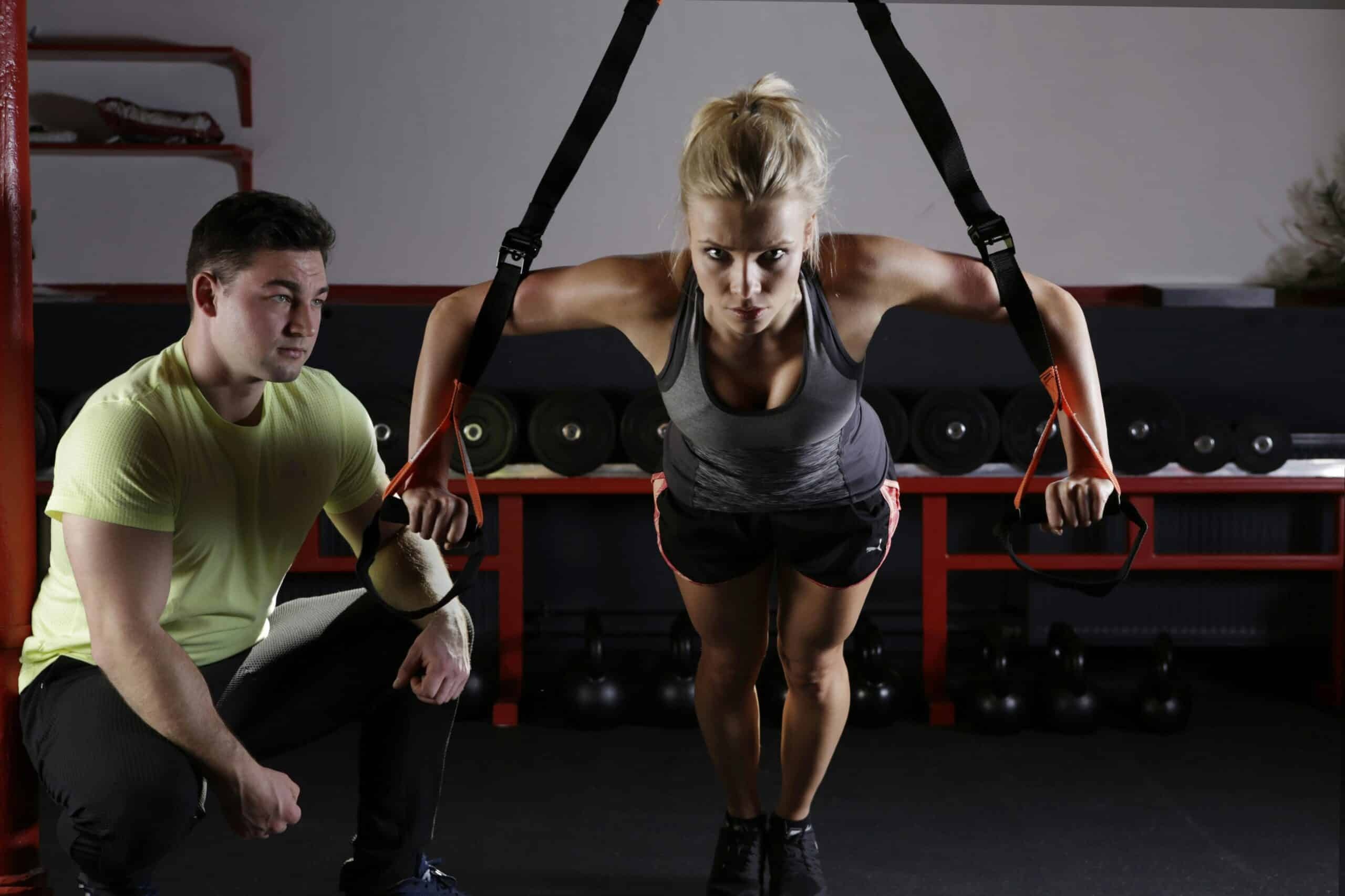 Woman performing TRX suspension training with a personal trainer assisting in a gym setting.